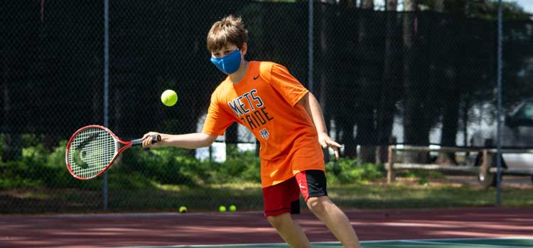 a kid playing tennis with a mask on at a summer camp that stayed open in 2020